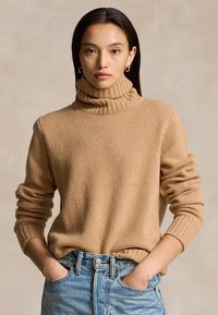 Woman with long dark hair wearing gold hoop earrings, a camel-colored turtleneck sweater, and light blue jeans with hands in pockets.