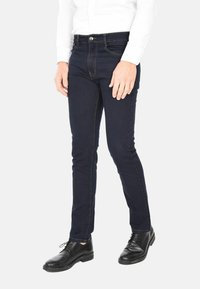 Uomo che indossa jeans slim-fit blu scuro, scarpe eleganti in pelle nera e una camicia bianca a maniche lunghe, in piedi contro uno sfondo semplice.