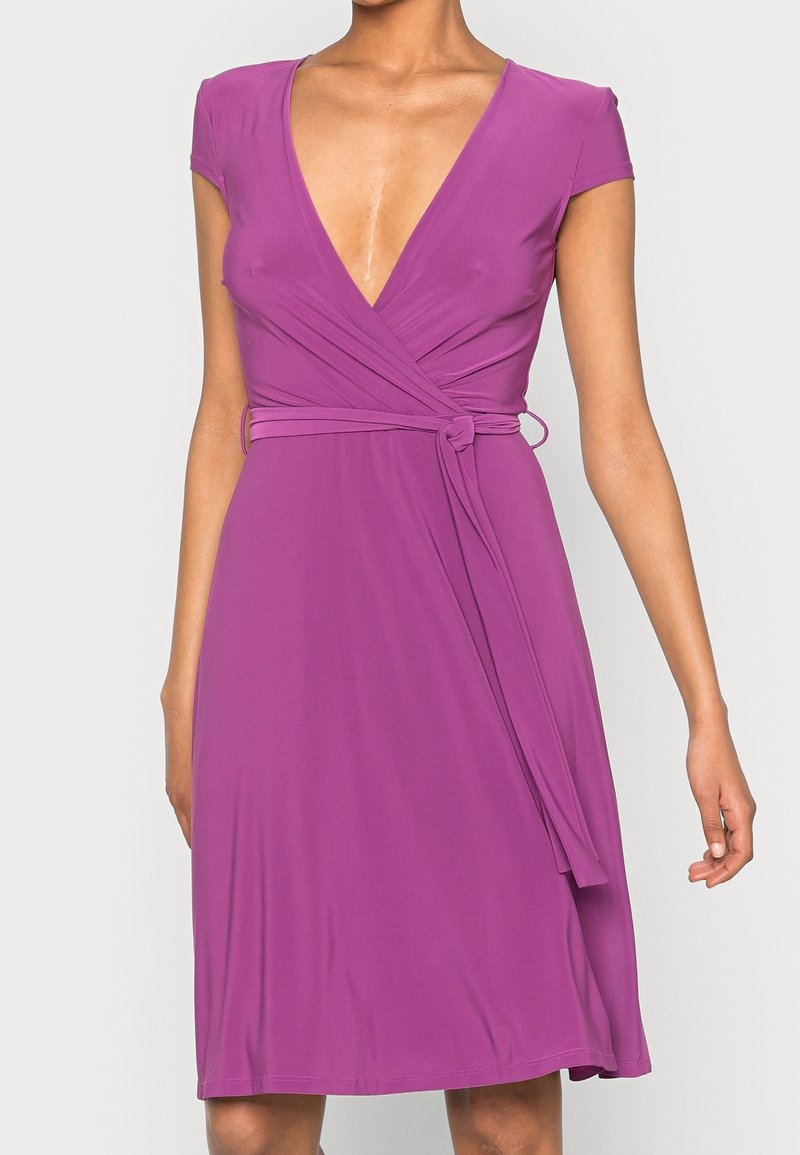 Robe portefeuille violette avec manches courtes, encolure en V et ceinture nouée à la taille, portée par une personne sur un fond uni.