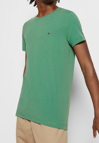 T-shirt vert à manches courtes en coton, avec un col rond et un petit logo rouge, blanc et bleu sur la poitrine.