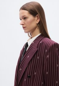 Burgundski suknjič z črtami in subtilnim vzorcem logotipa, dvoredni kroj, reverji z zarezo in teksturirana tkanina.