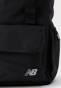 Mochila de tela negra con un bolsillo frontal con solapa y un logo plateado "NB". El diseño general es simple y cuenta con correas ajustables.