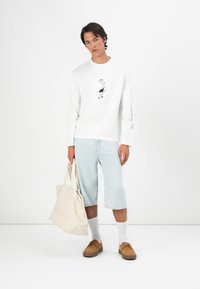 Witte lange mouwen shirt met grafisch detail, lichtblauwe knielange shorts, beige tas en bruine schoenen. Eenvoudige achtergrond, minimale vormgeving.
