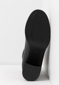 Semelle de chaussure noire avec grip texturé au talon, reposant contre un mur et un sol blancs, indiquant la taille 37 sur la semelle.