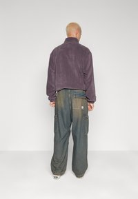 Fialová fleecová bunda s vysokým límcem, spárovaná s volnými, vybledlými modrými cargo kalhotami s bočními kapsami a detailem nášivky.