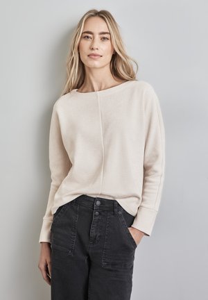 Beige geripptes Langarmoberteil mit weitem Ausschnitt und lockerer Passform, kombiniert mit hoch taillierten schwarzen Hosen mit Vordertaschen und Knopfverschluss.