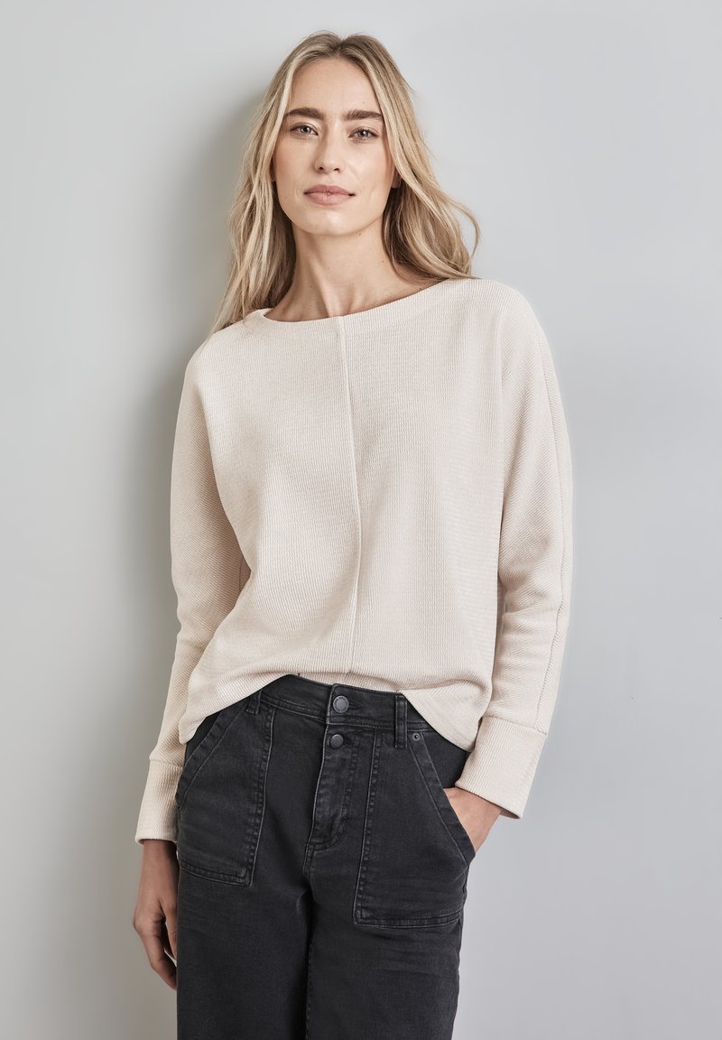 Beige geripptes Langarmoberteil mit weitem Ausschnitt und lockerer Passform, kombiniert mit hoch taillierten schwarzen Hosen mit Vordertaschen und Knopfverschluss.