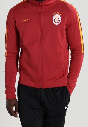 Veste de sport zippée rouge avec des rayures jaunes sur les manches, logo Galatasaray, swoosh Nike, portée avec un pantalon Nike noir.