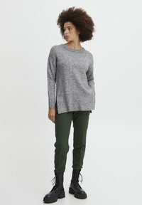 Pull gris avec ourlet côtelé et coutures d'épaule, associé à un pantalon vert fuselé et des bottes noires à lacets. Fond uni.