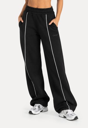 Pantalones deportivos negros de pierna ancha con ribetes verticales blancos, cintura elástica, bolsillos laterales, combinados con zapatillas plateadas y blancas.