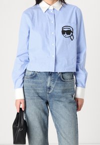 Kvinna klädd i en blå och vit randig skjorta med vit krage och manschetter, broderad ansikte på fickan, blå jeans, håller en svart handväska.