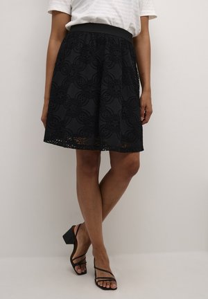 Frau trägt einen schwarzen knieumspielenden Rock aus Blumenspitze, ein weißes Oberteil und schwarze, mit Riemchen versehene Blockabsatzsandalen, steht mit gekreuzten Beinen.