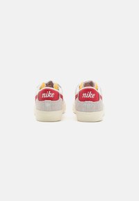 Sneaker in suede grigio con dettagli bianchi e rossi, caratterizzate da un design perforato e un prominente logo "nike" sul tallone. Suola color crema.