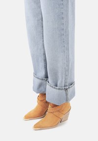 Botas de tobillo de ante color marrón con puntera afilada y detalles trenzados. Tacón cuña con acabado de grano de madera. Vaqueros cortos de color azul claro con los puños doblados.