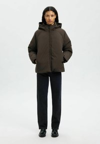Giacca a vento marrone con cappuccio, imbottita e con zip frontale e tasche laterali, indossata sopra pantaloni in denim scuro e scarpe nere, su uno sfondo chiaro.