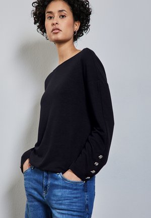 Maglietta nera a maniche lunghe in un tessuto morbido, con scollatura arrotondata e dettagli in bottone argentato sui polsini, abbinata a jeans blu.