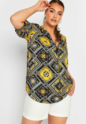 Blusa nera a maniche corte con motivo paisley giallo e bianco, con chiusura a bottoni e orlo curvo, abbinata a pantaloni corti bianchi.
