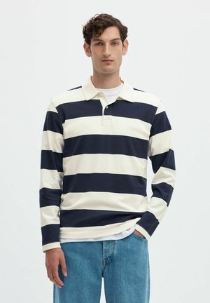 Homme aux cheveux bouclés et foncés portant un polo à manches longues à larges rayures blanches et bleu marine et un jean bleu, debout devant un fond uni.