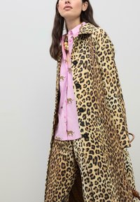 Leopard-mønstret frakke i tan og brun med sorte knapper, båret over en pink skjorte med små, brune dyremønstre.