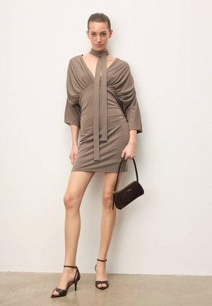 Femme debout contre un mur blanc, portant une mini-robe taupe froncée avec un détail de nouage long, tenant un petit sac à main marron foncé et des sandales à talons noires.