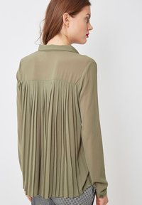 Blusa de manga larga verde oliva con un diseño de espalda plisada y un frente liso. Presenta un cuello y puños abotonados. Tejido texturizado.