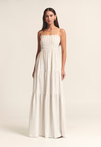 Maxi obleka z našitimi naborki na zgornjem delu, tankimi naramnicami in večplastno krilom. Iz lahkega blaga, v svetlo bež barvi z nežnimi črtami.