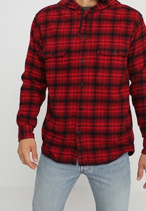 Muž nosící červenou a černou kostkovanou košili s kapucí a světle modré džíny, stojící na jednobarevném světlém pozadí.