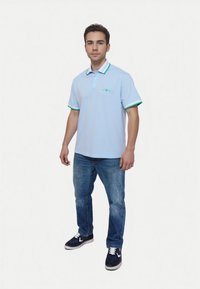 Hellblaues Poloshirt mit grün-weißen gestreiften Kragen und Ärmeln, kombiniert mit blauen Jeans und dunklen Sneakers. Verfügt über ein kleines Logo auf der Brust.