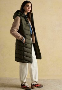 Kobieta w długiej, oliwkowej pikowanej kamizelce z kapturem, beżowej kurtce, białych spodniach i brązowych trampkach, stoi na beżowym tle.