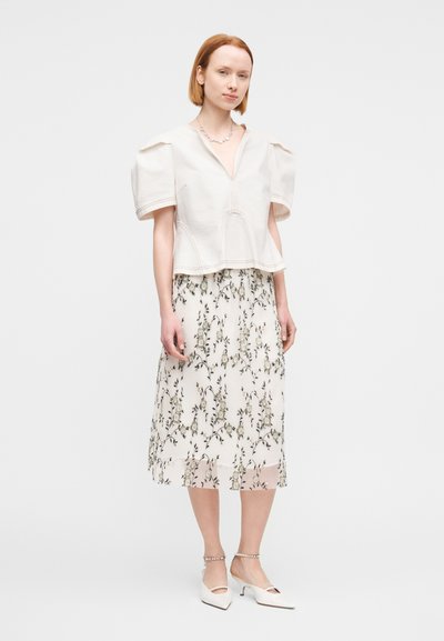 Άσπρο, κοντομάνικο τοπ με φουσκωτούς ώμους, σε συνδυασμό με φλοράλ, πλισέ φούστα με πράσινα και μπεζ μοτίβα. Άσπρα παπούτσια με τακούνι.