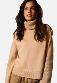 Maglione beige con collo alto e maniche e orlo a costine testurizzate. Design morbido e oversize, lunghezza cropped, adatto per essere indossato a strati.