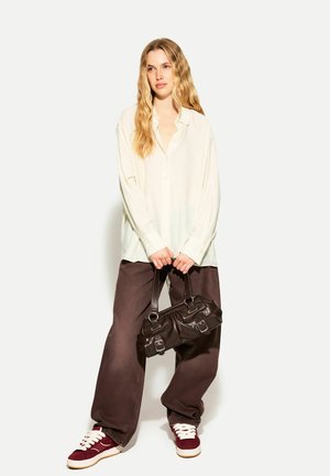 Übergroßes cremefarbenes Hemd mit Knöpfen, braune weitgeschnittene Hose und burgunderrote Sneaker, hält eine dunkelbraune Lederhandtasche mit metallischen Akzenten.