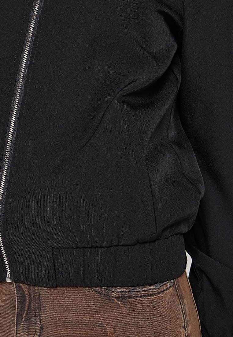Schwarze Bomberjacke mit glatter Textur, Frontreißverschluss und elastischem Saum sowie Bündchen, kombiniert mit braunen strukturierten Hosen.