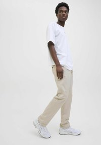 Jeune homme debout en pantalon beige, t-shirt blanc ample et baskets blanches sur un fond blanc uni.