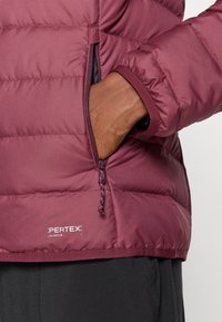 Κάπα μωβ με φερμουάρ στο πλάι, ελαστικούς καρπούς και γυαλιστερή, καπιτονέ υφή. Περιλαμβάνει την επωνυμία "Pertex Quantum".