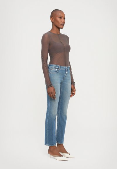 Transparante bruine langemouwen top met een knoopsluiting en gerimpelde details, gecombineerd met lichtblauwe rechte jeans en witte hakken.