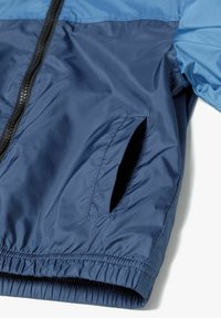 Chaqueta rompevientos de color azul marino con una textura ligera y suave, que cuenta con dos bolsillos laterales y un dobladillo elástico, complementada por una sección superior de un azul más claro.