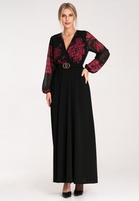 Czarna maksiodzienna sukienka z przezroczystą koronką w kwiatowy wzór, długimi rękawami i ściągniętą talią z akcentem w postaci podwójnego pierścienia. 