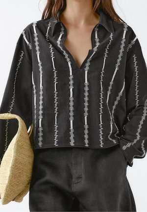 Black collared shirt with white linear embroidery, short sleeves, and a loose fit. Paired with a woven straw bag.
