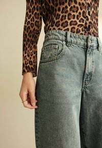 Išblukę mėlyni plačios kojos džinsai su penkių kišenių dizainu ir matomomis siūlemis, derinami su rudais leopardų rašto marškiniais, turinčiais raukinėtą tekstūrą ir mygtukus.