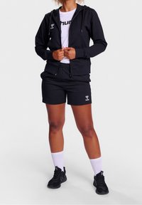 Sudadera negra con cremallera y bolsillo frontal, combinada con pantalones cortos deportivos negros. Logo blanco en la sudadera y en los pantalones cortos. Zapatillas negras y calcetines blancos.