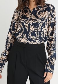 Námořnická blůza s růžovým květinovým vzorem, zapínání na knoflíky vpředu, dlouhé rukávy a jemná látka; v kombinaci s černými kalhotami s vysokým pasem.