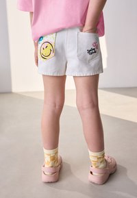Shorts di denim bianchi con toppe di faccine sorridenti colorate sulle tasche posteriori. Abbinati a Crocs rosa e calzini con motivo floreale giallo.