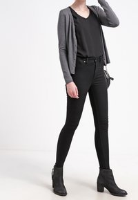 Cardigan gris, top negro de escote en pico, jeans ajustados negros y botas de tobillo. El cardigan es de punto; los jeans son ceñidos; las botas tienen un tacón cuadrado.