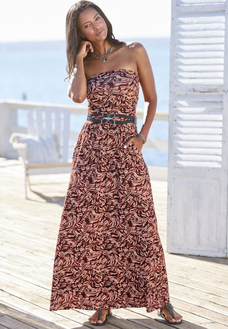 Trägerloses Maxikleid mit einem roten und cremefarbenen Blättermuster, tailliert mit einem schwarzen Gürtel. Leichter Stoff, getragen mit Flip-Flops.