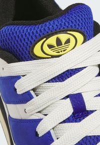 Blue and white athletic shoe featuring mesh fabric, contrasting accents, thick white laces, and a yellow retro logo badge.