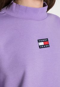 Sudadera púrpura con cuello alto, que presenta un logotipo bordado en negro "TOMMY JEANS" junto a un diseño rectangular en rojo y blanco.