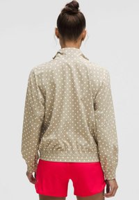 Jachetă bej cu buline, cu talie strânsă, manșete elastice și guler, asociată cu pantaloni scurți roz aprins. Textură netedă, material ușor.