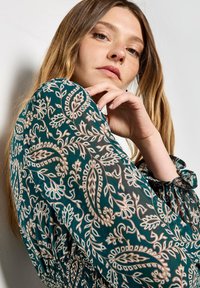 Blusa de gasa teal con intrincados patrones de paisley en blanco y dorado, que presenta un puño con fruncido y detalle de cintura fruncida.