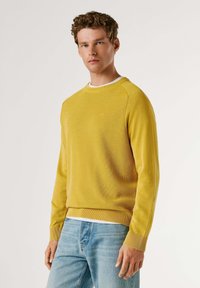 Gele gebreide trui met geribbelde manchetten en zoom, voorzien van een ronde halslijn en een gestructureerde stof. Draagt over lichtblauwe spijkerbroek.